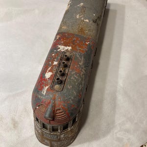May include: A vintage toy train engine made of metal with red, white, and gray paint. The engine has a detailed design with a rounded front and a long, rectangular body. The engine has a series of small holes on the top, likely for air vents. The engine is weathered and shows signs of age and use.