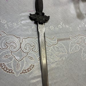 May include: An antique dagger with a detailed, ornate design. The handle is dark, possibly black, with a textured grip. The blade is silver with etched patterns. The crossguard features a decorative winged design. The dagger is displayed on a patterned surface.