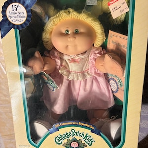 Op de afbeelding: Een Cabbage Patch Kids 15th Anniversary Special Edition pop met geel garen haar, groene ogen en een roze gingham jurk met een witte kraag. De pop zit in de originele verpakking, inclusief een geboortecertificaat en adoptiepapieren.