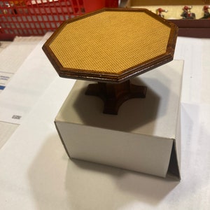 Può includere: Un tavolo ottagonale in miniatura con una base in legno marrone e un piano intrecciato. Il tavolo è appoggiato su una scatola bianca.