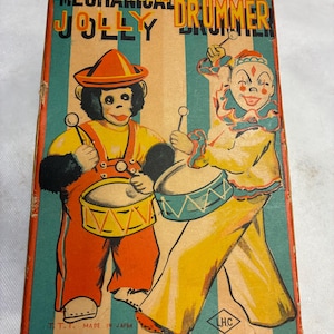 以下が含まれることがあります： ヴィンテージ「Mechanical Jolly Drummer」おもちゃ箱。箱には、ドラムを演奏する猿とピエロのイラストが描かれています。箱はオレンジ、黄色、ティール色で、上部に「Mechanical Jolly Drummer」の文字があります。箱には「Made in Japan」と記載されています。