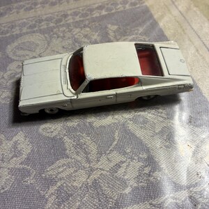 May include: A white die-cast toy car featuring a bright red interior, visible windows, and a trunk lid. The exterior paint shows signs of wear and chipping, particularly along the roofline and edges of the metal body of this vintage vehicle.