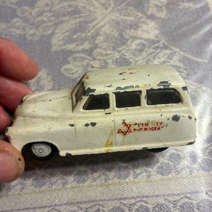 May include: A vintage white die-cast toy ambulance with black windows and wheels. The side features a red Star of David symbol, Hebrew text, and the words RED MAGEN. The paint shows significant wear, with gray metal visible along the edges.