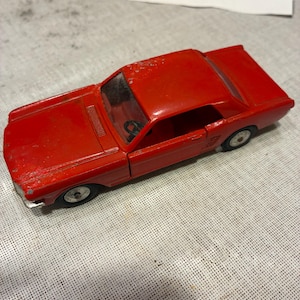 May include: A vintage red die-cast toy car featuring a two-door coupe body style, a clear plastic windshield, a black steering wheel visible inside the cabin, and silver-colored wheels. The red paint shows signs of wear and minor chipping.