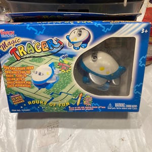 May include: A blue and white toy airplane with a magic marker pen. The toy is part of a game called "Magic Tracer" and includes 16 plastic jigsaw pieces. The box is blue and white with the words "Magic Tracer" and "Hours of Fun" printed on it. The toy is for ages 3 and up.