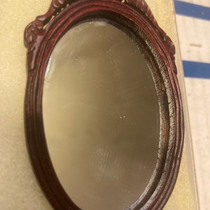 May include: A small, oval-shaped mirror with a dark wood frame. The frame has intricate carvings and a decorative top.