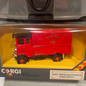 May include: A red toy truck with a black cab and a white "Royal Mail" sign on the side. The truck has a black grill and black wheels. The truck is in a box with a yellow background and the words "Corgi Classics" and "AEC 508 forward control 5ton Cabover" printed on it.