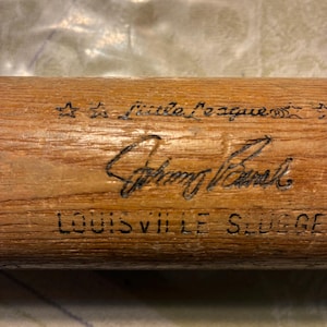 May include: A light brown wooden baseball bat featuring black printed text and graphics. The markings include the words Little League, a signature, and the text LOUISVILLE SLUGGER. The wood grain is visible across the surface of the bat.