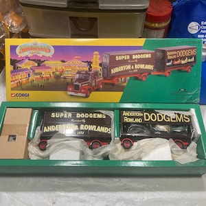 May include: A Corgi Classics model of a vintage truck hauling a trailer with a sign that reads "Super Dodgems Presented By Anderton & Rowlands Est. 1854". The truck is brown with red accents and the trailer is black with white lettering. The model is in a green box with white foam padding.