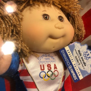 May include: A Cabbage Patch Kid doll dressed in a blue and red gymnastics leotard with the USA Olympic logo and the words "Olympikids 1996 Official Team Moscow" on the front. The doll has brown hair and brown eyes. The doll is holding a tag that says "USA Gymnastics Olympikids Special Edition Molly Joanelle was born in the Cabbage Patch on October 15 14598-0903".