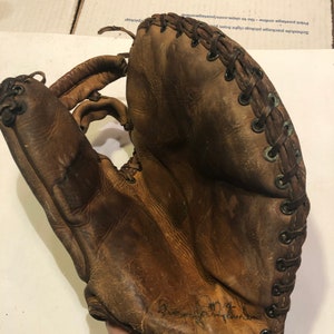May include: A brown leather baseball glove with laces and a signature on the palm. The glove is worn and has a vintage look.