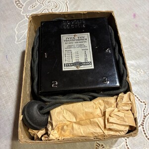 Könnte beinhalten: Ein Vintage Ives Spielzeugtransformator, Modell Nr. 204-100 Watt, in seiner originalen Pappschachtel. Der schwarze Transformator hat ein Etikett mit Spannungs- und Verdrahtungsanweisungen. Die Box enthält ein schwarzes Kabel und einen schwarzen, runden Stecker.