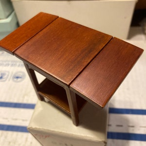 May include: A small, wooden table with a drop-leaf design. The table has a brown finish and a shelf underneath.