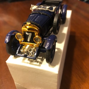 Puede incluir: Un coche de juguete vintage azul marino con detalles dorados, incluyendo la parrilla y los faros. El número 7 se muestra en la parte delantera. El coche tiene ruedas negras y se exhibe en una caja blanca.