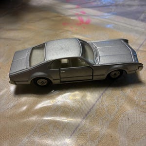 May include: A silver-colored die-cast toy car featuring a two-door coupe body style, clear windows, and black wheels. The vehicle has a metallic finish, a defined hood, a roofline, and a trunk section, showcasing a classic automotive design.