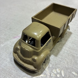 May include: A vintage toy dump truck in a muted olive green color. The truck features a detailed front grill, open cab, and a rectangular cargo bed. The number "943957" is visible on the side of the cab. This classic toy truck is a nostalgic collectible.