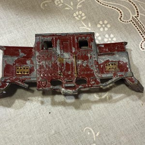 Puede incluir: Un vagón de tren de juguete antiguo con un aspecto desgastado. El vagón es principalmente rojo con áreas de metal gris expuesto, pequeñas ventanas y detalles dorados. El texto "No. 1" es visible en la parte delantera.