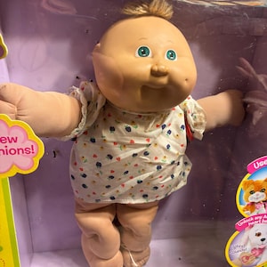 Op de afbeelding: Een Cabbage Patch Kids pop met een rond gezicht, groene ogen en een klein plukje bruin haar. De pop draagt een wit shirt met korte mouwen en een klein blauw en geel bloemenpatroon. De pop zit verpakt in een paarse doos met tekst en afbeeldingen.