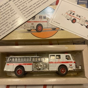 Könnte beinhalten: Ein weißes Spielzeugfeuerwehrauto mit roten Akzenten und einer Leiter. Das Auto hat die Aufschriften "DENVER FIRE RESCUE" und "DENVER" an der Seite. Das Auto befindet sich in einer Schachtel mit weißem Hintergrund.