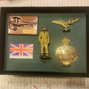 May include: A display case containing a collection of miniature items, including a gold aeroplane, a gold eagle, a gold medal with a crown, a miniature figure of a pilot in a khaki uniform, and a small Union Jack flag with the text "Charles Rumney Samson".