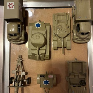 May include: A collection of vintage toy military vehicles in shades of tan and olive green. The set includes trucks, tanks, a jeep, and artillery, each bearing Israeli symbols. The toys are arranged on a light brown surface.