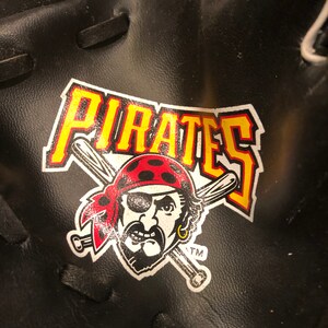 Puede incluir: Guante de béisbol de cuero negro con un logotipo de "Pirates" amarillo y rojo que presenta un pirata de dibujos animados con un parche en el ojo y un arete. El logotipo también incluye bates de béisbol cruzados.