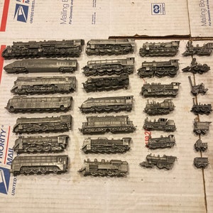 May include: A collection of 18 miniature pewter train engines. The engines are all different styles and sizes, and some have details like smoke stacks and wheels. The engines are arranged in rows on a brown cardboard surface.