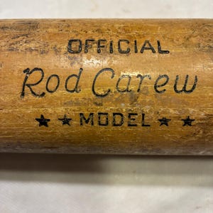 May include: Close-up of a wooden baseball bat with the text "OFFICIAL Rod Carew MODEL" in black lettering. The bat has a light brown color with visible wood grain and some wear.