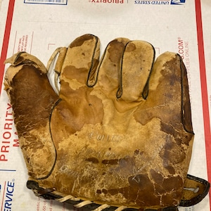 May include: A brown leather baseball glove with a black stitching detail. The glove is worn and has a faded appearance. The glove is signed "K. Lewis" in white ink.