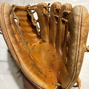 May include: A well-worn, tan leather baseball glove. The glove has visible stitching, lacing, and the text "Regent 711 Signature" is printed on the palm. The glove shows signs of use and wear.