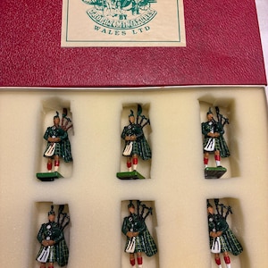 May include: A collection of six miniature bagpiper figures in a red box. The figures are dressed in green plaid kilts and jackets, with white sporrans and red socks. The box lid features a label that reads "Traditional Models for the Collector".