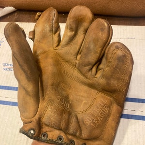 May include: A well-worn, tan leather softball glove. The glove has visible wear and tear, with stitching and lacing details. The words "SOFT BALL LEAGUE" are embossed on the glove. The glove is held up by a person.