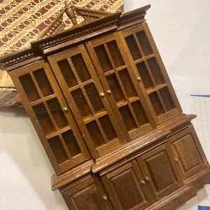 May include: A wooden dollhouse hutch with glass doors and a bottom cabinet. The hutch has a decorative top and is made of a dark brown wood.