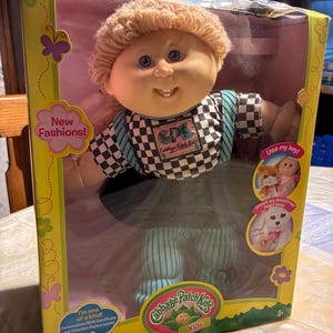 Puede incluir: Una muñeca Cabbage Patch Kids con cabello de lana rubio y ojos azules, vistiendo una camisa a cuadros blancos y negros y pantalones a rayas verde azulado. La muñeca está dentro de una caja de cartón amarilla y morada con texto.