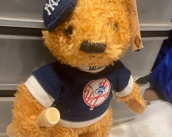 May include: A brown teddy bear wearing a blue and white New York Yankees jersey and a blue baseball cap with the NY logo. The bear is holding a wooden baseball bat.
