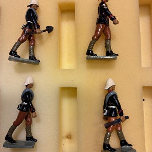 Puede incluir: Cuatro soldados de juguete en miniatura en una vitrina. Cada soldado viste una chaqueta negra, pantalones marrones y un casco blanco. Uno sostiene una pala, otro un rifle. Las figuras están sobre bases grises.