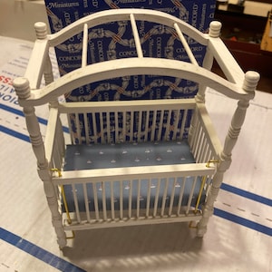 May include: A white miniature dollhouse crib with a canopy. The crib has a blue floral patterned mattress and gold-colored accents. A box labeled "CONCORD Miniatures" is in the background.
