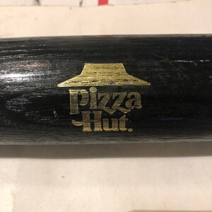 May include: A black wooden baseball bat with a gold Pizza Hut logo. The logo features the iconic red roof and the words "Pizza Hut" in gold lettering.