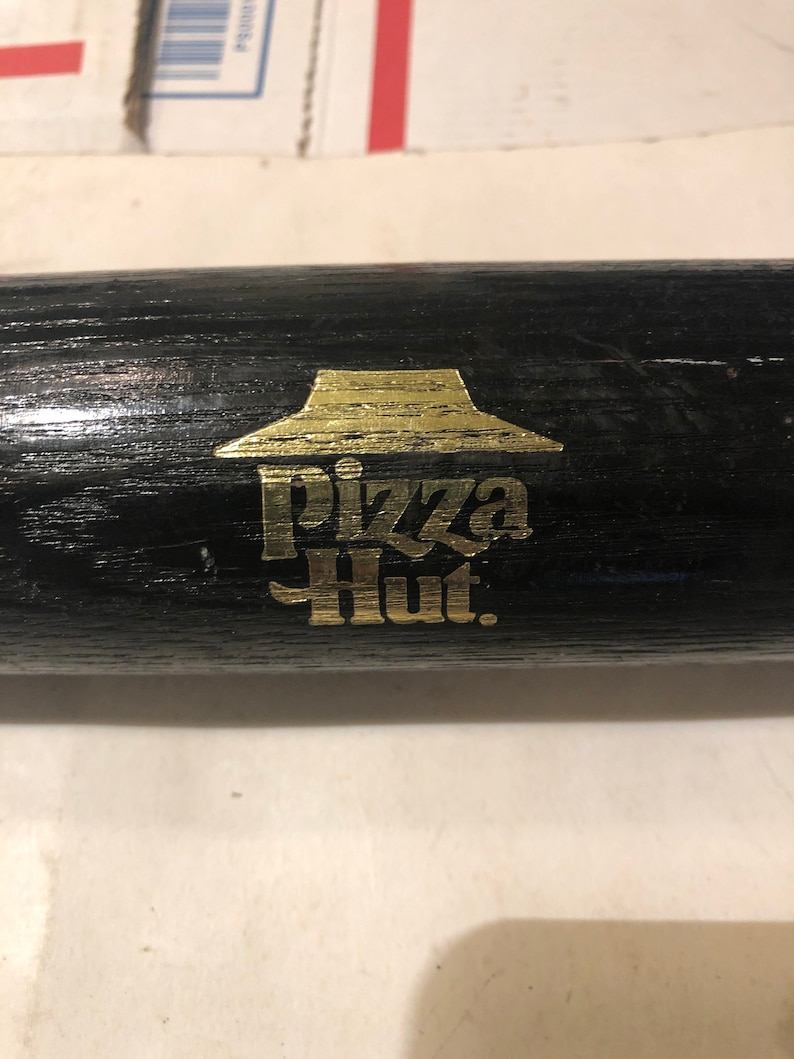 May include: A black wooden baseball bat with a gold Pizza Hut logo. The logo features the iconic red roof and the words "Pizza Hut" in gold lettering.