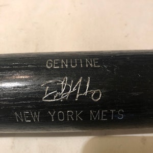May include: A black baseball bat with the text "GENUINE" and "NEW YORK METS" in silver lettering. The bat is signed with a silver signature that reads "Edgardo Alfonzo".