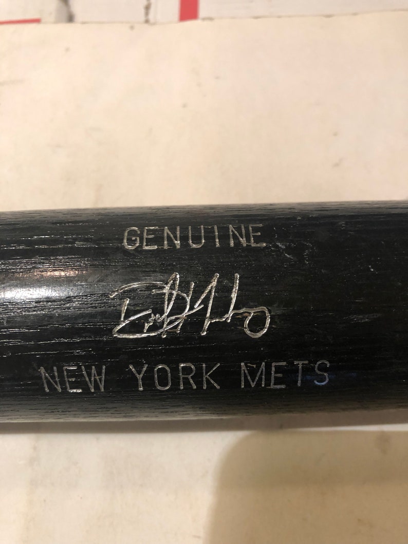 May include: A black baseball bat with the text "GENUINE" and "NEW YORK METS" in silver lettering. The bat is signed with a silver signature that reads "Edgardo Alfonzo".