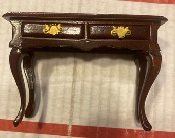 May include: A dark brown wooden table with two drawers and ornate gold handles. The table has curved legs and a simple design.