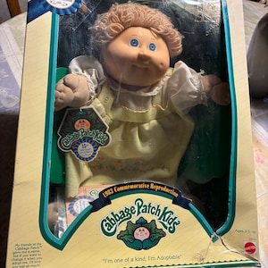 Op de afbeelding: Een Cabbage Patch Kids 15th Anniversary Special Edition pop in de originele verpakking. De pop heeft krullend blond haar, blauwe ogen en draagt een gele jurk met witte mouwen. De doos bevat de tekst 1983 Commemorative Reproduction.