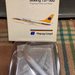 May include: A 1:500 scale model of a Boeing 737-500 airplane in the colors of Hapag-Lloyd. The model is made of metal and is displayed in a clear plastic case.