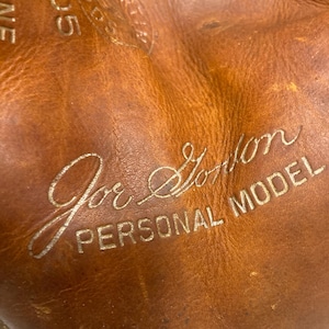 May include: Close-up of a well-worn, brown leather baseball glove. The glove features the embossed text "Joe Gordon PERSONAL MODEL" in silver lettering. The leather shows signs of use, with visible creases and wear.