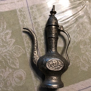 May include: A silver metal teapot with intricate engraved designs and Arabic script. The teapot has a spout, a handle, and a lid.