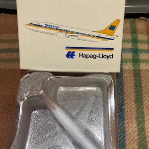 May include: A white and yellow Boeing 737-400 airplane model with the text "Boeing 737-400" and "Flugzeugmodell aus Metall" on the box. The airplane model is in a clear plastic display case. The airplane has the text "Hapag-Lloyd" on the side.