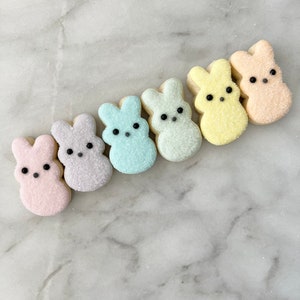 May include: Six pastel-colored bunny-shaped cookies decorated with black eyes and sugar sprinkles. The cookies are arranged in a row on a white marble surface.