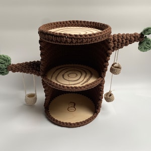 Può includere: Un tronco d'albero in crochet marrone con tre ripiani e due cesti appesi. Il tronco d'albero ha un ramo in crochet verde con foglie su entrambi i lati.