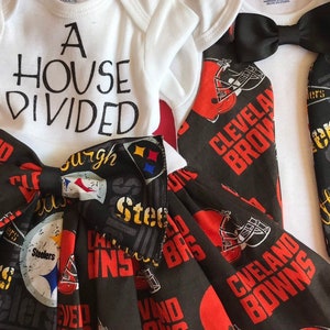 May include: A white onesie with the text "A House Divided" and a black bow tie. A black and red skirt with the text "Cleveland Browns" and "Pittsburgh Steelers" repeated on the fabric.